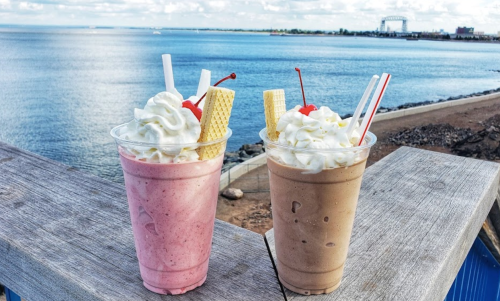 Two milkshakes, one pink and one brown, topped with whipped cream, cherries, and wafers, by a scenic waterfront.
