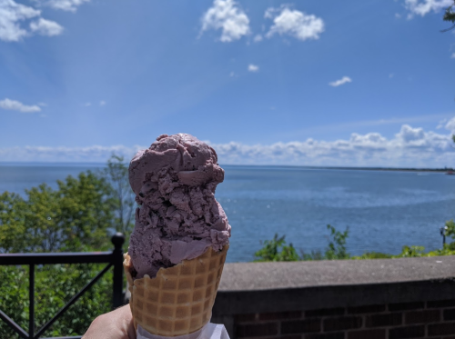 A hand holds a cone of purple ice cream against a scenic lake and blue sky with fluffy clouds.