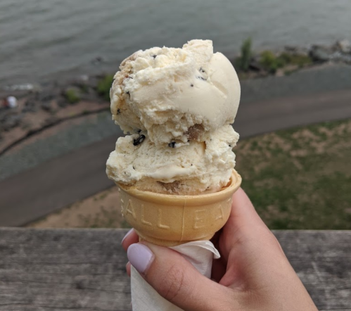 A hand holding a cone with two scoops of ice cream by a body of water, with a scenic view in the background.