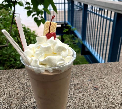 A frosty milkshake topped with whipped cream, a cherry, and a wafer, set against a scenic outdoor background.