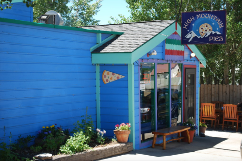 Bright blue building with a sign for "High Mountain Pies," featuring a pizza slice decoration and outdoor seating.