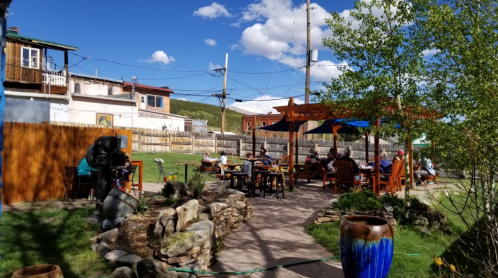 A sunny outdoor patio with people dining, surrounded by greenery and rustic decor, under a blue sky with clouds.