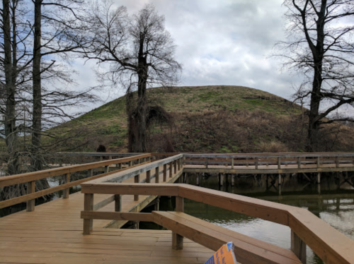 A wooden walkway leads to a grassy hill, surrounded by trees and water under a cloudy sky.
