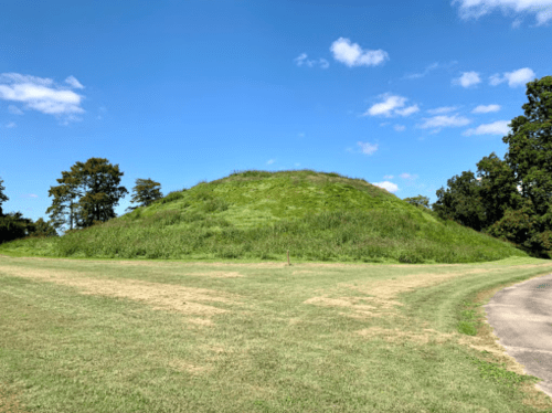 A grassy mound surrounded by trees under a clear blue sky, with a path curving around the base.