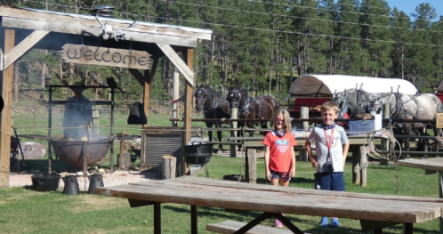 Two children stand smiling in front of a rustic welcome sign, with horses and a wagon in the background.