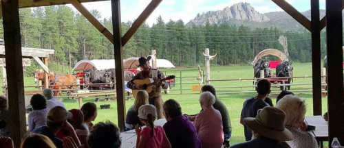 A performer plays guitar for an audience outdoors, with horses and mountains in the background.