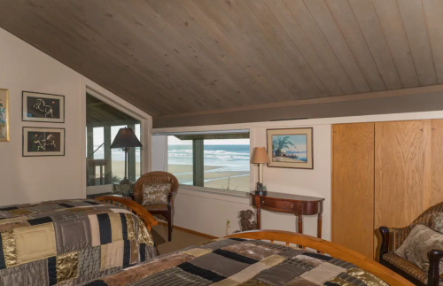 Cozy bedroom with two beds, a lamp, and a view of the beach through large windows.