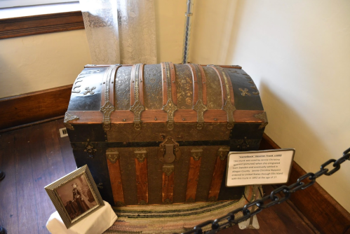 A vintage trunk with metal accents, displayed beside a framed photo and an informational plaque in a museum setting.