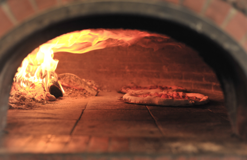 A wood-fired oven with flames, baking pizzas on a stone surface.