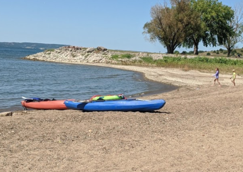 Two kayaks on a sandy beach by a calm lake, with trees in the background and two children walking along the shore.