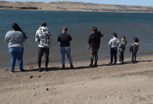 A group of people standing by a lake, facing the water, with hills in the background on a sunny day.