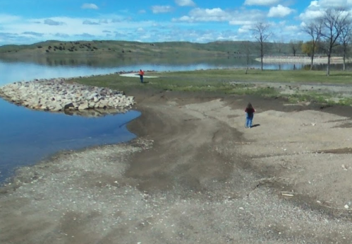A sandy shoreline with a person walking along the water's edge and another person in the distance by the lake.
