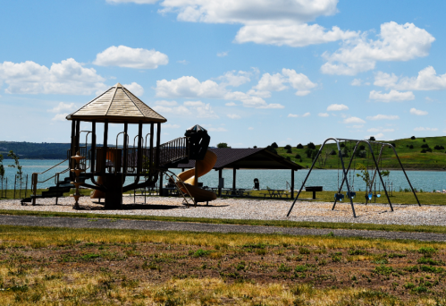A playground with a wooden structure and swings near a lake, under a blue sky with fluffy clouds.