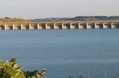 A concrete dam spans a calm blue lake, surrounded by green hills under a clear sky.