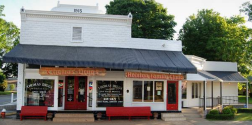 Historic drugstore building with red doors and signage, surrounded by greenery, showcasing early 20th-century architecture.