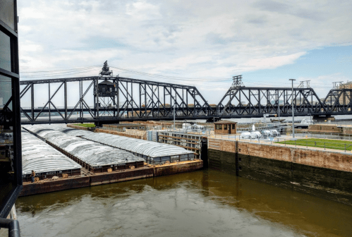 A view of a river lock with a barge and a lift bridge in the background under a cloudy sky.