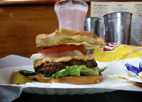 A juicy burger with lettuce, tomato, onion, and sauce, served with pickles and a milkshake in the background.