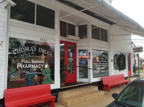 Exterior of Thomas Drugs pharmacy, featuring red benches and large windows displaying services and ice cream offerings.