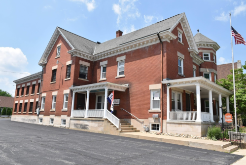 Historic brick building with a porch, flagpole, and landscaped front, set against a clear blue sky.