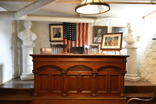 A wooden desk in a historic room with busts, framed portraits, and an American flag in the background.