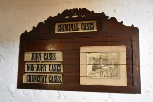 A wooden board displaying categories for court cases: Criminal, Jury, Non-Jury, and Chancery, with a historical photo.