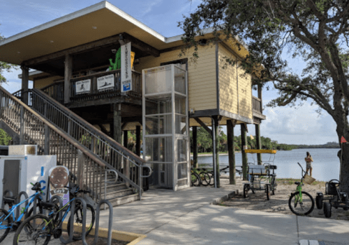 A two-story building by a lake with an elevator, surrounded by bicycles and picnic areas under a clear sky.