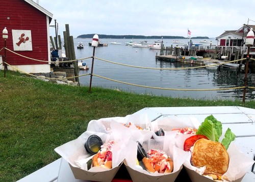 A scenic view of a harbor with lobster rolls in the foreground, boats in the water, and a red building in the background.