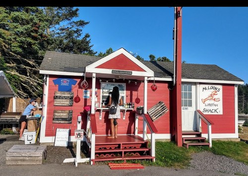 A red seafood shack with a woman ordering at the window, surrounded by trees and outdoor seating.
