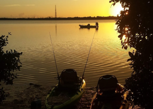 Two kayaks on the shore at sunset, with a boat in the distance and a calm water surface reflecting the golden light.