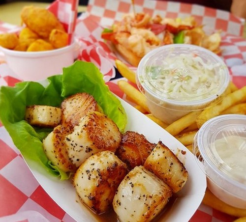 A plate of grilled scallops on lettuce, with sides of coleslaw, fries, and shrimp, all on a checkered tablecloth.