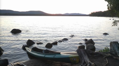 A kayak rests on a rocky shore at sunset, with calm water and distant mountains in the background.