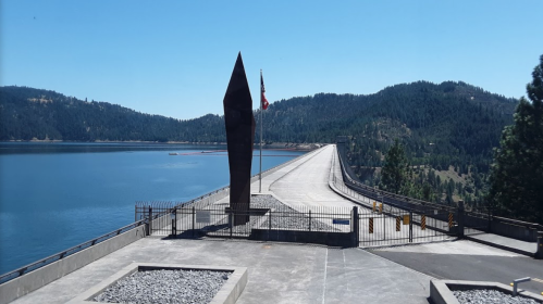 A dam with a tall monument, surrounded by water and mountains under a clear blue sky.