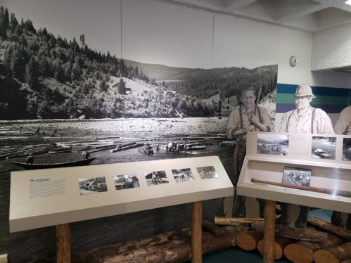 Black and white mural of a river scene with historical figures, alongside display panels and wooden logs in a museum setting.