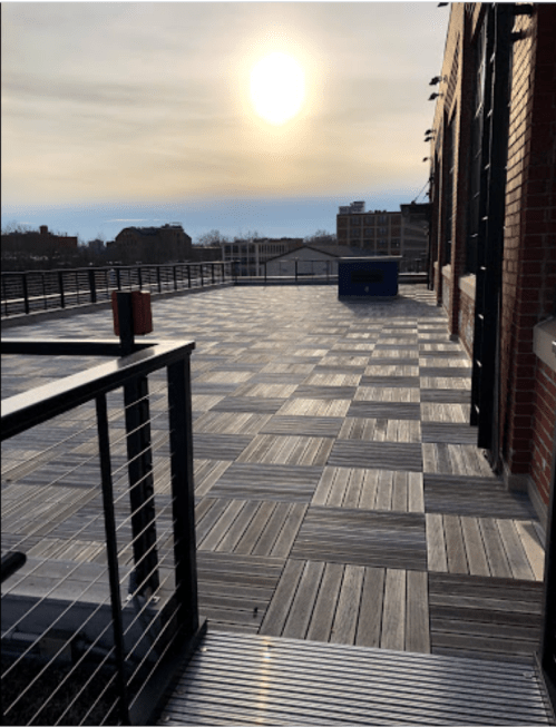 A sunlit rooftop terrace with a wooden deck and a railing, overlooking a cityscape in the distance.