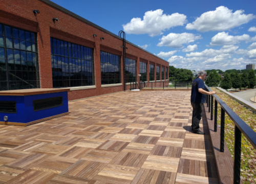 A man stands on a wooden deck overlooking a scenic view with blue skies and clouds in the background.