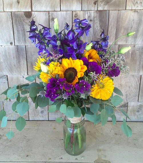 A vibrant bouquet of sunflowers, purple flowers, and greenery in a glass jar against a rustic wooden background.