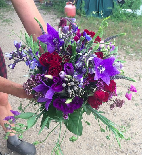 A vibrant bouquet of purple and red flowers, including roses and lilies, held by a person outdoors.