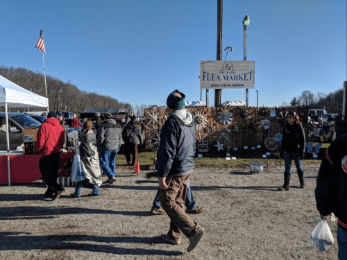 A busy flea market scene with people walking, tents, and a sign for the market in the background.