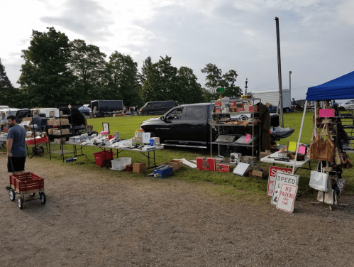A bustling outdoor market with tables of various items for sale, surrounded by green grass and parked vehicles.
