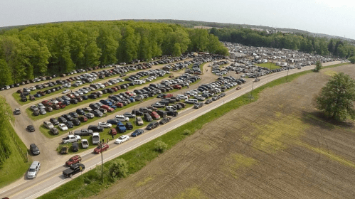 Aerial view of a large parking lot filled with cars, surrounded by trees and fields.