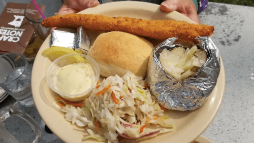 A plate with a bread roll, fried fish fillet, coleslaw, a baked potato in foil, and a small cup of sauce.