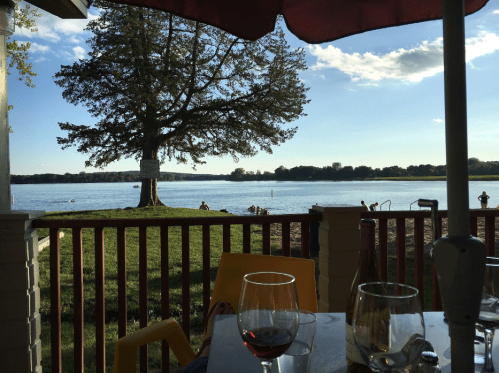 A serene lakeside view with a tree, people swimming, and glasses of wine on a table in the foreground.