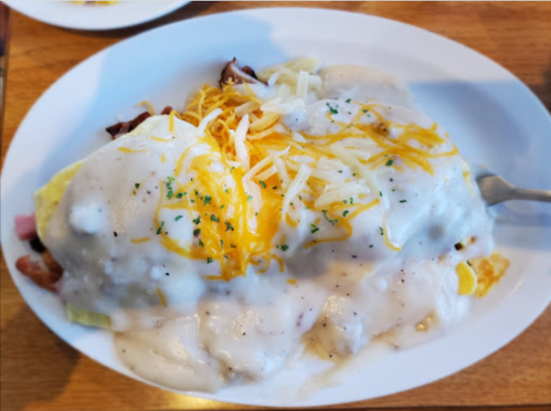 A plate of omelette topped with creamy gravy, cheese, and herbs, served with a side of bacon and ham.