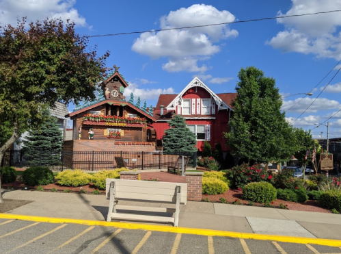A charming red house with decorative flowers, a clock tower, and a landscaped yard under a blue sky with fluffy clouds.