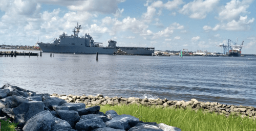 A large military ship docked near a waterfront, with boats and cranes in the background under a partly cloudy sky.