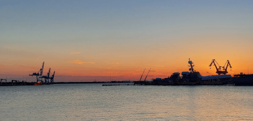 Sunset over a calm harbor, with silhouettes of cranes and ships against a colorful sky.