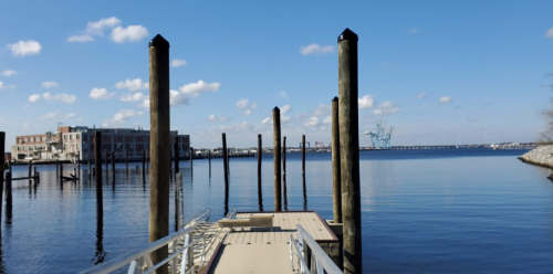 A calm waterfront scene featuring a wooden dock with tall posts, under a clear blue sky and distant industrial structures.