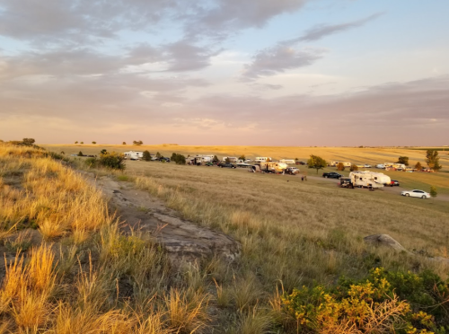 A grassy hillside with RVs and cars parked in a field under a colorful sunset sky.