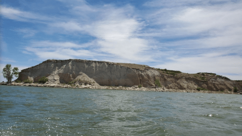 A rocky cliff rises above a calm river under a blue sky with scattered clouds. Green trees line the water's edge.