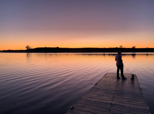 A person stands on a dock at sunset, holding a dog, with calm water reflecting the colorful sky.